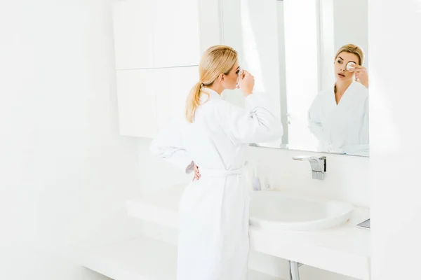 Selective focus of attractive and blonde woman in white bathrobe using cotton pad and looking at mirror — Stock Photo