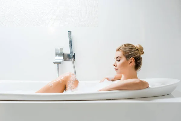Hermosa y rubia mujer tomando baño con espuma y mirando hacia otro lado en el baño - foto de stock