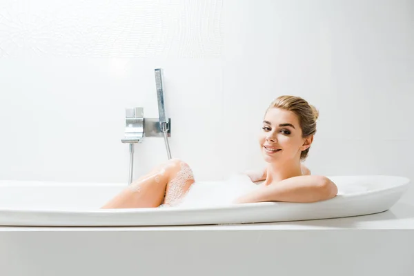 Attraktive und blonde Frau, die im Badezimmer mit Schaum badet und in die Kamera schaut — Stockfoto
