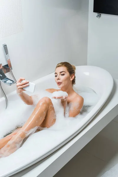 Привлекательная и улыбающаяся женщина принимает ванну, дует в пену и делает селфи в ванной комнате — стоковое фото