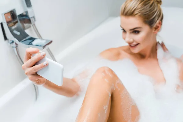 Messa a fuoco selettiva di attraente e sorridente donna fare il bagno con schiuma e prendere selfie in bagno — Foto stock