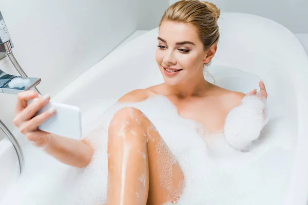 Foco seletivo de mulher atraente e sorridente tomando banho e tomando selfie no banheiro — Fotografia de Stock