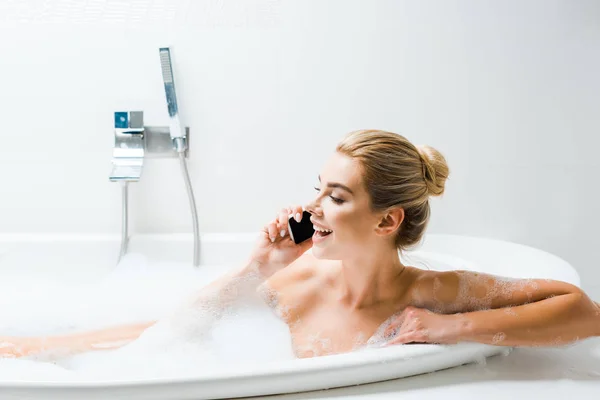 Mujer atractiva y sonriente tomando baño con espuma y hablando en el teléfono inteligente en el baño - foto de stock