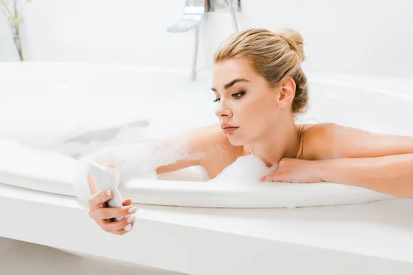 Attraktive und blonde Frau, die im Bad mit Schaum badet und das Smartphone benutzt — Stockfoto