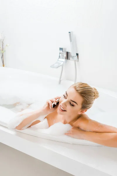 Bela e sorridente mulher tomando banho com espuma e falando no smartphone no banheiro — Fotografia de Stock