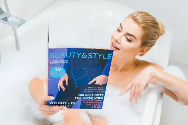 Вибірковий фокус привабливої і блондинки, приймаючи ванну з піною і читаючи журнал краси і стилю у ванній — стокове фото