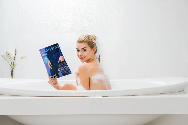 Atractiva y sonriente mujer tomando baño con espuma y la celebración de la belleza y el estilo de la revista en el baño - foto de stock