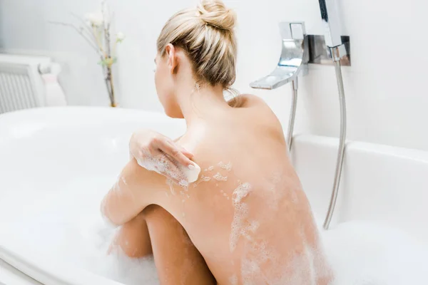 Vista trasera de mujer rubia tomando baño con espuma y sosteniendo jabón en el baño - foto de stock