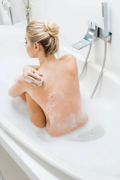 Vista trasera de mujer rubia tomando baño con espuma y sosteniendo jabón en el baño - foto de stock