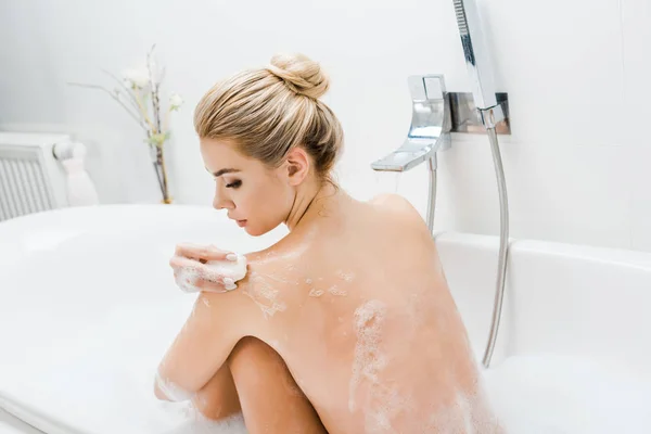 Schöne und blonde Frau nimmt Bad mit Schaum und hält Seife im Badezimmer — Stockfoto