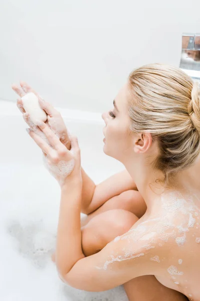 Attractive and blonde woman taking bath with foam and holding soap in bathroom — Stock Photo