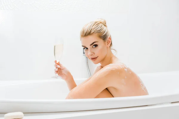 Hermosa y rubia mujer tomando baño y sosteniendo copa de champán en el baño - foto de stock