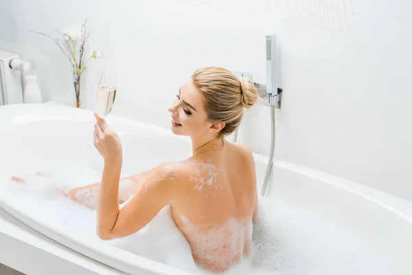 Schöne und lächelnde Frau badet und hält Champagnerglas im Badezimmer — Stockfoto