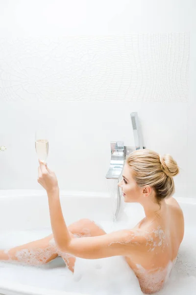 Schöne und lächelnde Frau badet und hält Champagnerglas im Badezimmer — Stockfoto