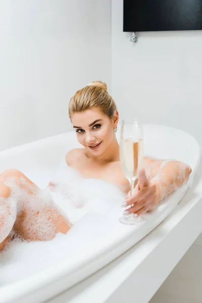 Foco seletivo da mulher bonita e sorridente tomando banho e segurando copo de champanhe no banheiro — Fotografia de Stock