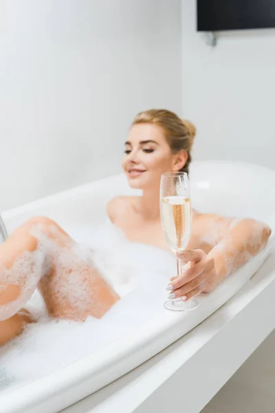 Selective focus of champagne glass holding by beautiful and smiling woman in bathroom — Stock Photo