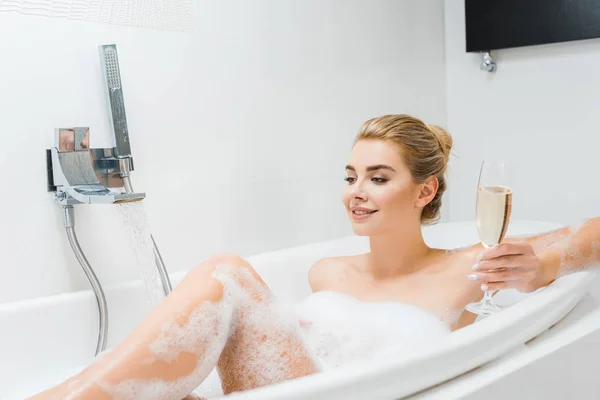 Bela e sorridente mulher tomando banho e segurando taça de champanhe no banheiro — Fotografia de Stock