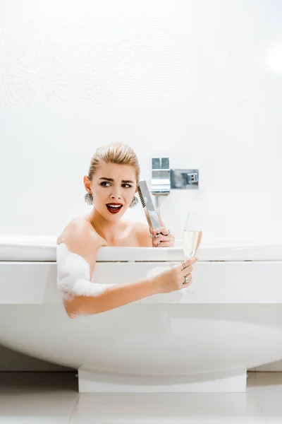 Hermosa y rubia mujer tomando baño, sosteniendo copa de champán y hablando en el baño - foto de stock