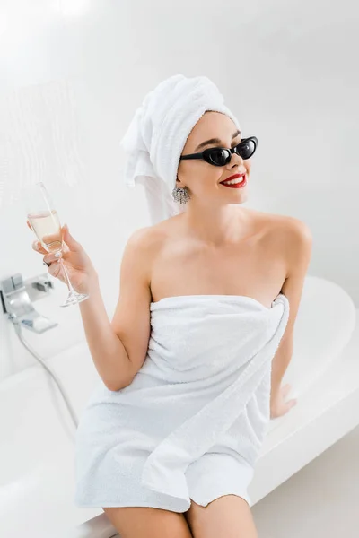 Mulher atraente e sorridente em óculos de sol e toalhas segurando copo de champanhe no banheiro — Fotografia de Stock