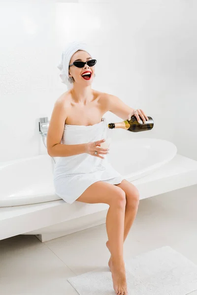 Beautiful and smiling woman in sunglasses and towels holding champagne glass and bottle — Stock Photo