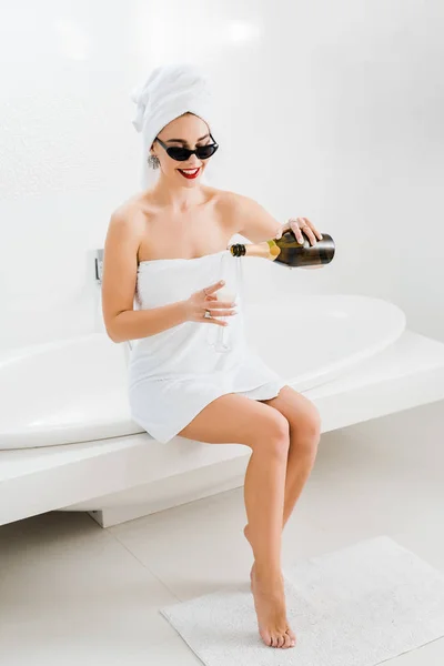 Mujer atractiva y sonriente en gafas de sol y toallas sosteniendo copa de champán y botella - foto de stock