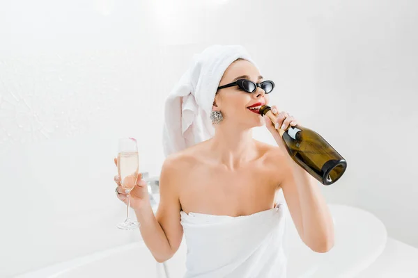 Atractiva mujer en gafas de sol y toallas sosteniendo copa de champán y beber de la botella — Stock Photo