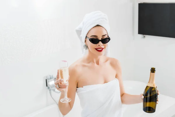 Mujer atractiva y feliz en gafas de sol y toallas celebración de copa de champán y botella — Stock Photo