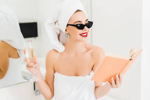 Attractive and smiling woman in sunglasses and towels holding champagne glass reading book — Stock Photo
