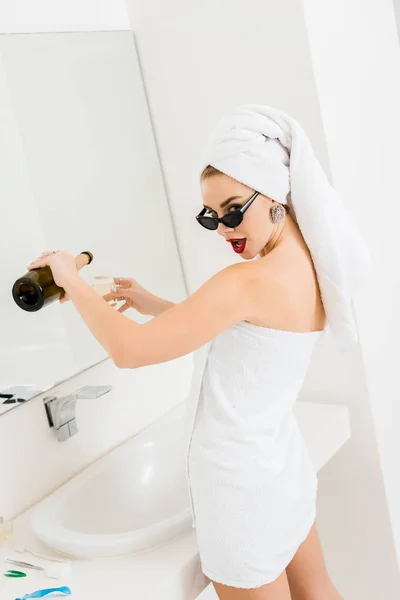 Attraente e sorridente donna in occhiali da sole e asciugamani in possesso di bicchiere di champagne e bottiglia e guardando la fotocamera — Foto stock