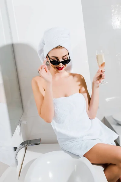 Attractive and smiling woman in sunglasses and towels holding champagne glass in bathroom — Stock Photo