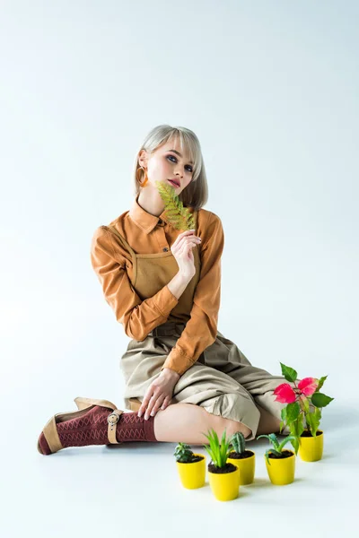 Beautiful stylish girl posing with fern leaf near flower pots on white — Stock Photo