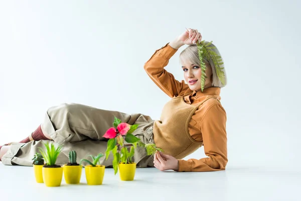 Hermosa chica con estilo mirando a la cámara y sosteniendo hojas de helecho mientras posa cerca de macetas aisladas en blanco - foto de stock