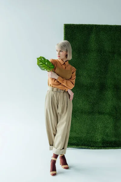 Bela jovem segurando folha monstera enquanto posando no branco com grama verde — Fotografia de Stock