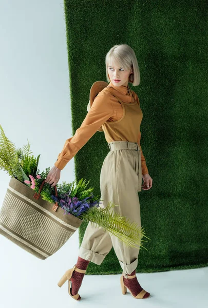Hermosa mujer joven con estilo sosteniendo bolsa con helecho y flores en blanco con hierba verde - foto de stock