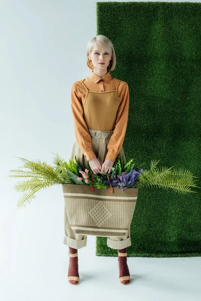 Schöne stilvolle Mädchen blickt in die Kamera, während sie Tasche mit Farn und Blumen auf weiß mit grünem Gras hält — Stockfoto