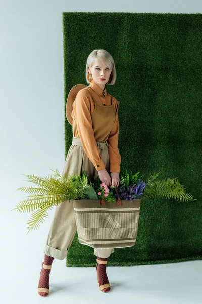 Beautiful stylish girl looking at camera while holding bag with fern and flowers on white with green grass — Stock Photo