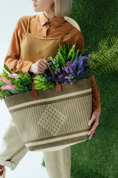 Abgeschnittene Ansicht einer stilvollen jungen Frau mit Tasche mit Farn und Blumen auf weißem mit grünem Gras — Stockfoto