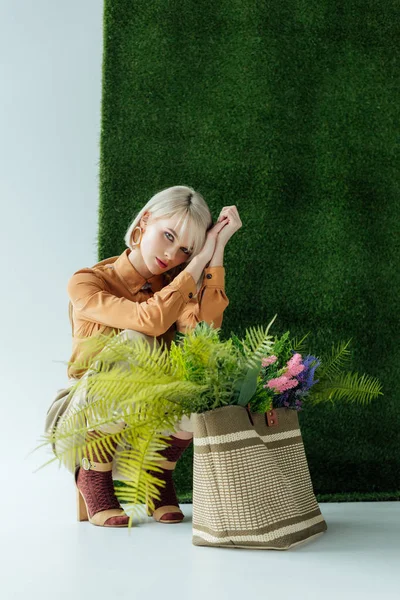 Beautiful fashionable girl posing near bag with fern and flowers on white with green grass — Stock Photo