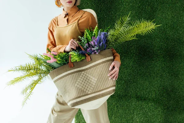 Partial view of stylish young woman holding bag with fern and flowers on white with green grass — Stock Photo