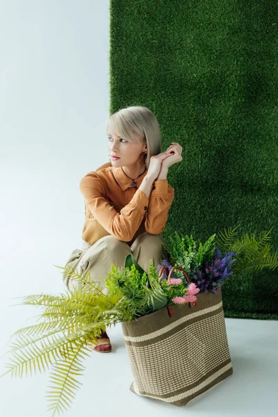 Beautiful stylish girl posing near bag with fern and flowers on white with green grass — Stock Photo