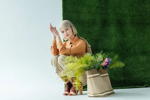 Красива модна дівчина позує біля сумки з папороті і квітами на білому з зеленою травою — стокове фото