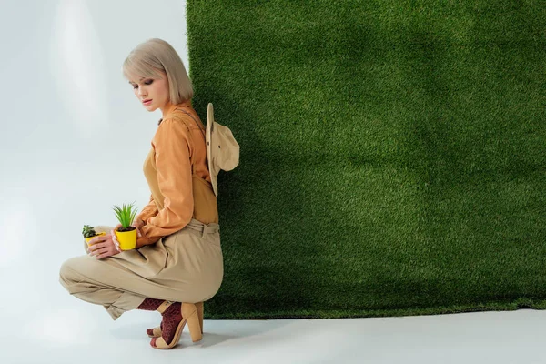 Красивая стильная девушка сидит и держит цветочные горшки на сером с зеленой травой — стоковое фото