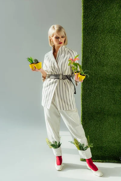 Belle fille élégante posant tout en tenant des pots de fleurs sur gris avec de l'herbe verte — Photo de stock