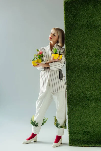 Hermosa chica elegante posando mientras sostiene macetas en gris con hierba verde - foto de stock