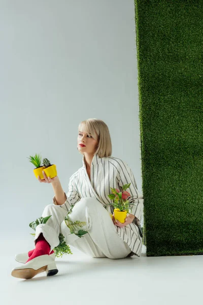 Красивая стильная девушка смотрит в сторону, держа цветочные горшки на сером с зеленой травой — стоковое фото
