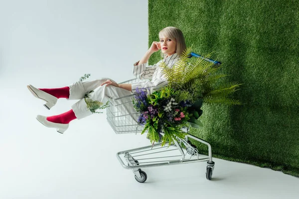 Schönes stilvolles Mädchen, das in die Kamera schaut, während es im Einkaufswagen mit Farn und Blumen auf weißem mit grünem Gras sitzt — Stockfoto