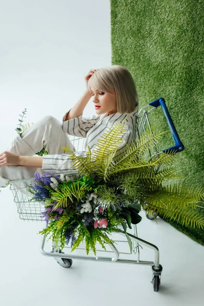 Belle fille élégante assise dans le panier avec fougère et fleurs sur blanc avec herbe verte — Photo de stock