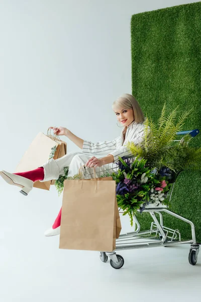 Schönes stylisches Mädchen blickt in die Kamera, während es im Einkaufswagen mit Farn, Blumen und Einkaufstaschen auf weiß mit grünem Gras sitzt — Stockfoto