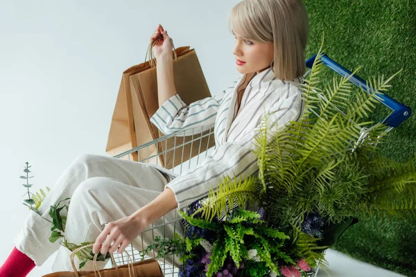 Hermosa chica elegante sentado en el carro con helecho, flores y bolsas de compras en blanco con hierba verde - foto de stock