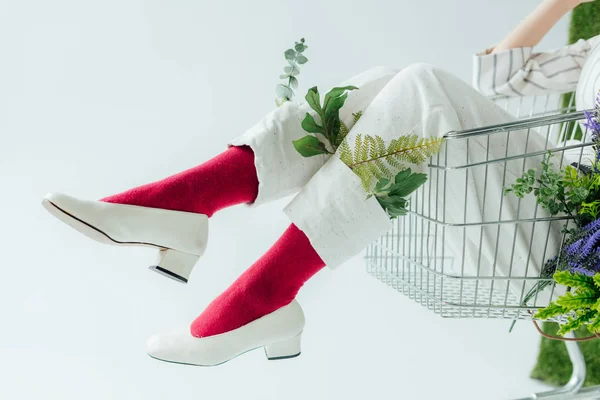 Abgeschnittene Ansicht einer Frau in weißer Hose mit grünen Blättern und stilvollen Schuhen, die in einem Einkaufswagen sitzt, isoliert auf weiß — Stockfoto